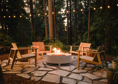 Teak modern Adirondack-styled chairs around a Rook Fire Table with string lights hanging overhead amongst tall evergreen trees